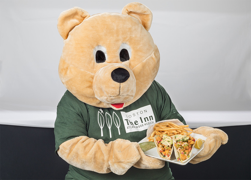 Hope the Bear, the mascot, from The Inn at Longwood, holding a plate of fish tacos from The Longwood Grille and Bar